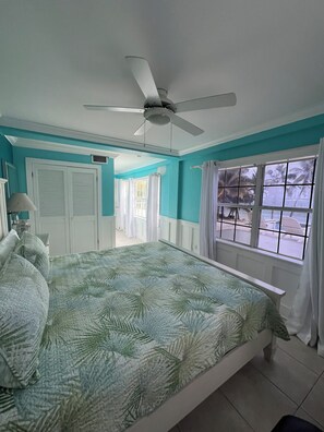 A bedroom with a bed, a ceiling fan, a window with curtains, and a view of the outside. - Snuggle Beach  Romantic & Charming 1-bedroom Apt. in enchanting East End with AC (East End)