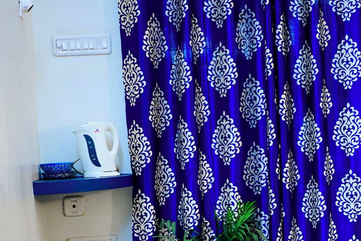 Ein Blick in einen Raum mit einem blauen, gemusterten Vorhang, einem weißen Wasserkocher, einer weißen Tasse und einem Blumentopf.