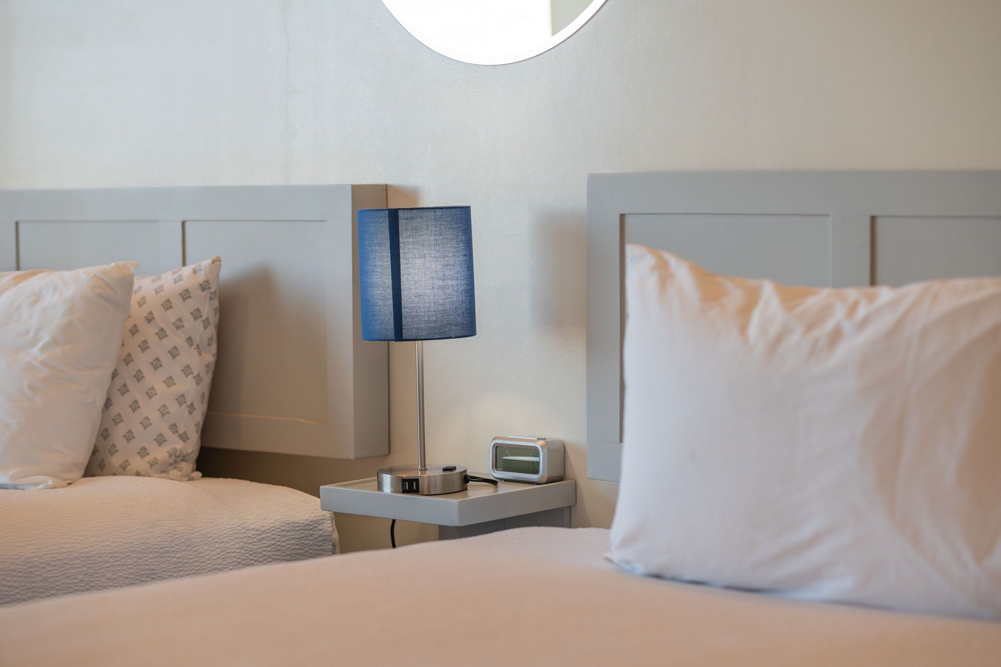Een hotelkamer met twee bedden, een nachtkastje met een lamp en een digitale klok, en een ronde spiegel aan de muur.