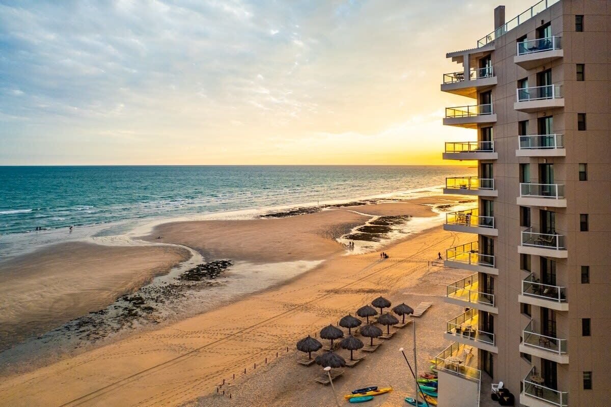 A beachfront hotel with a view of the ocean, a sandy beach, and a row of umbrellas.