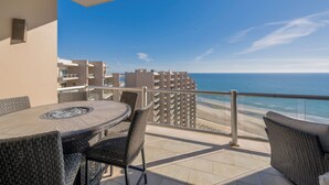 Een balkon met een ronde tafel en rieten stoelen, met uitzicht op een strand en een helderblauwe lucht.