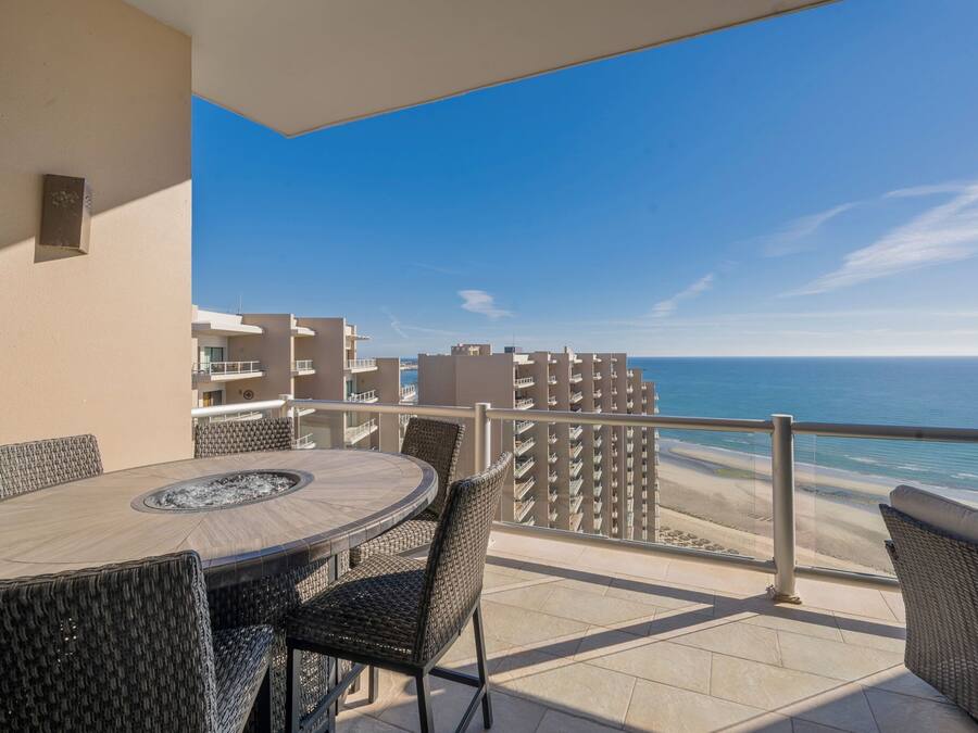 A balcony with a round table and wicker chairs, overlooking a beach and a clear blue sky.