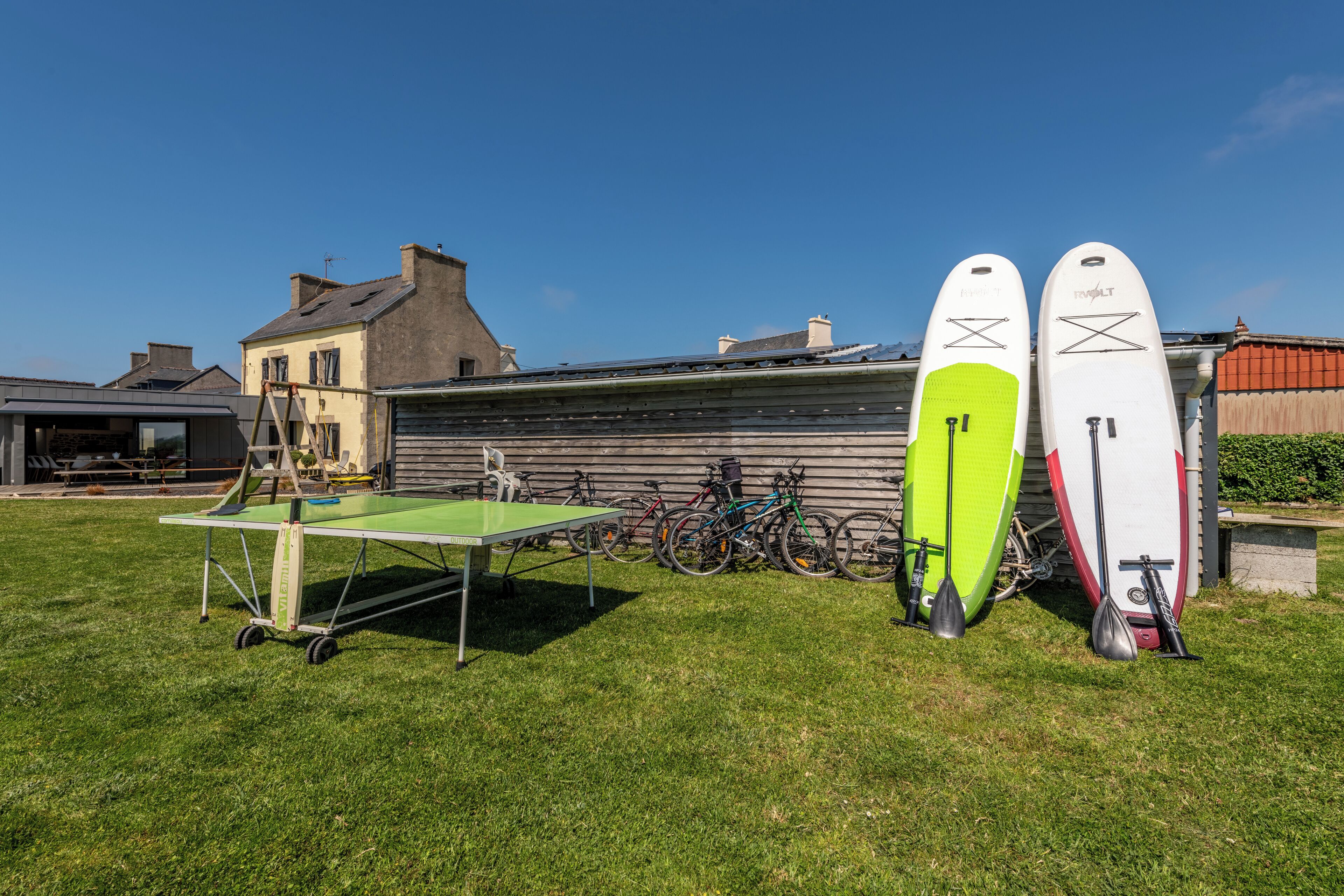 Ett grönområde utomhus med ett bordtennisbord, cyklar och surfbrädor.