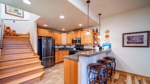 A kitchen with wooden cabinets, a stainless steel refrigerator, a microwave, a stove, and a sink. There are three stools at the island. - Black Bear 50 - Ski-In/Out Durango Townhome (Durango)