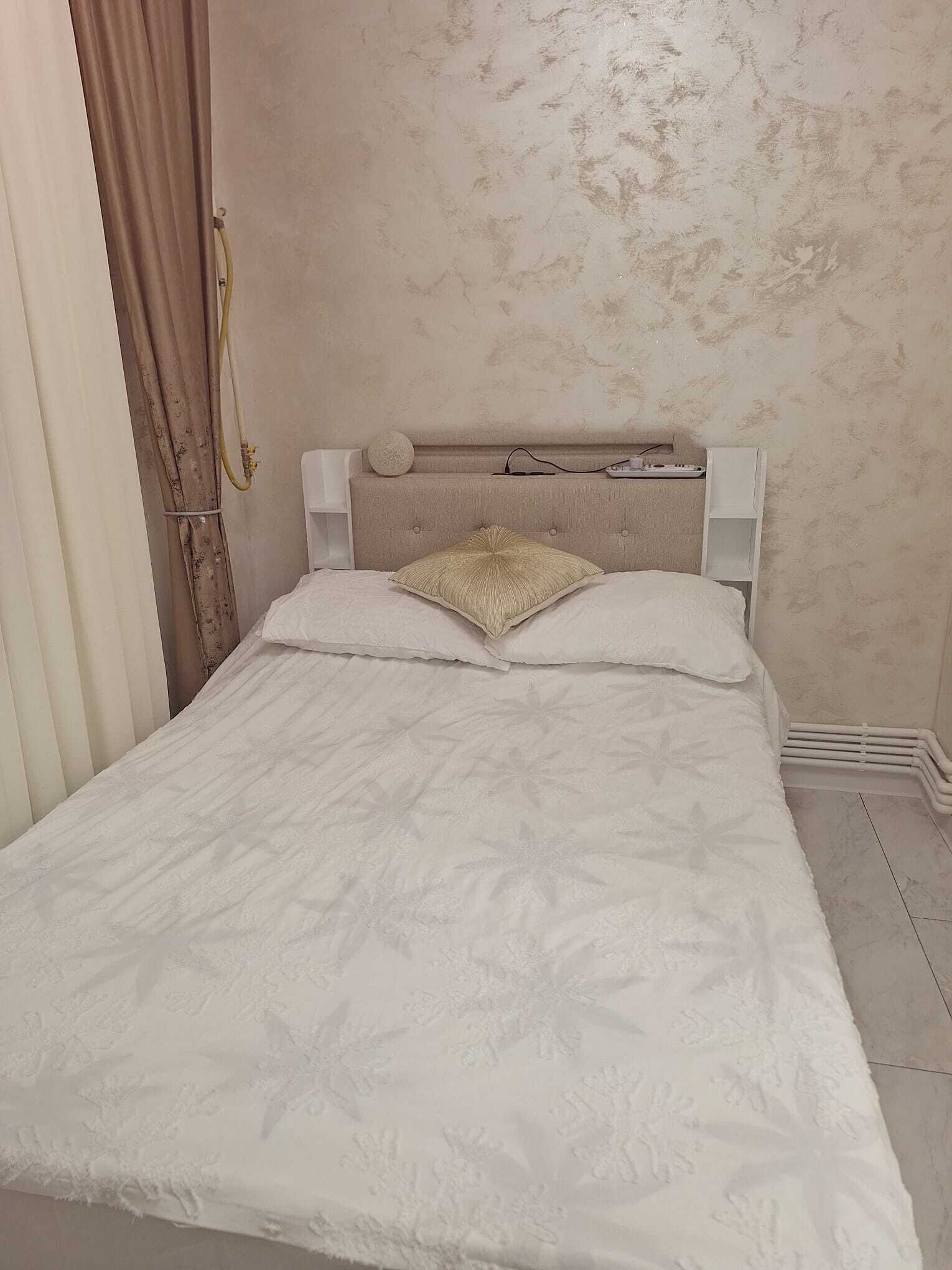 A bed with a white bedspread and two pillows, a headboard with a round object, and a white bedside table with a power outlet.