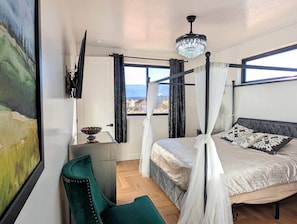 Et soveværelse med himmelseng, en grøn lænestol, et natbord med lampe, og udsigt til det fri gennem et vindue.
