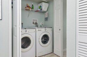 A laundry room with two front-loading washing machines, a shelf above with cleaning supplies, and a door leading to another room. -  Treasure Island condo with pool and jacuzzi (Treasure Island)