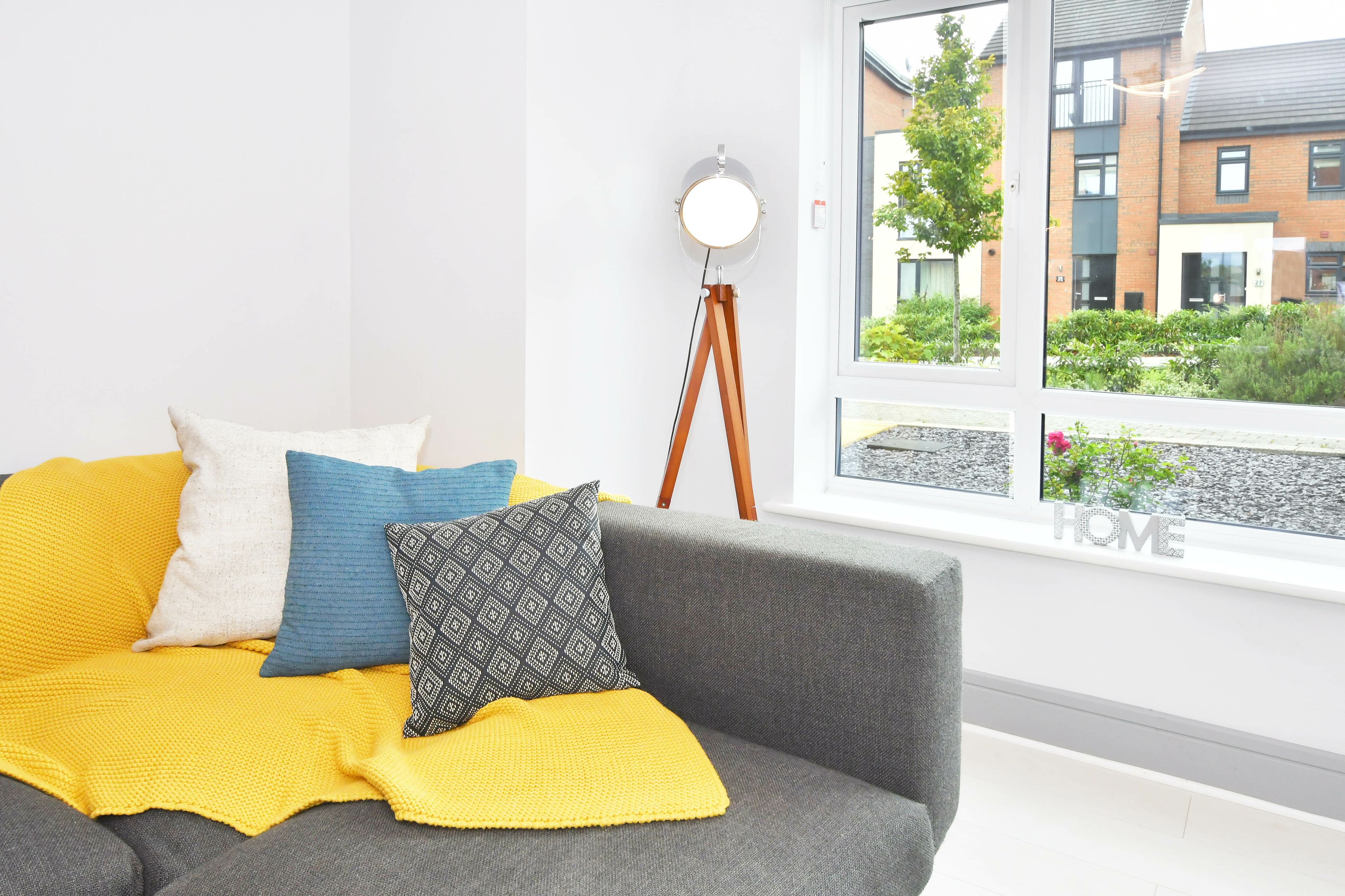 Ein modernes Wohnzimmer mit einem grauen Sofa, einer gelben Strickdecke und gemusterten Kissen. Eine Stehlampe steht an der Wand, und ein Fenster bietet Blick auf ein Gebäude und Grünflächen.