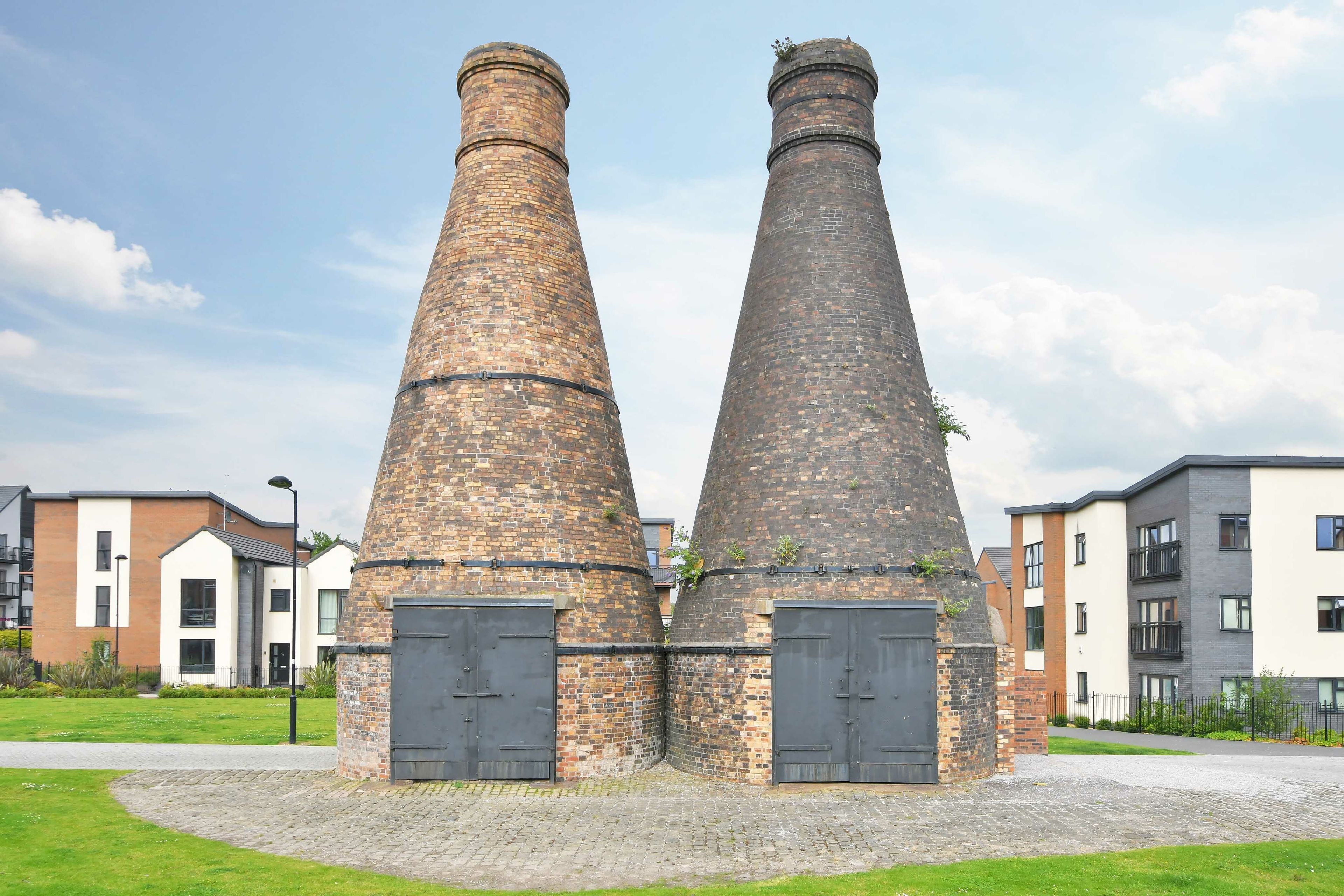 Two brick chimneys with doors, surrounded by modern buildings.