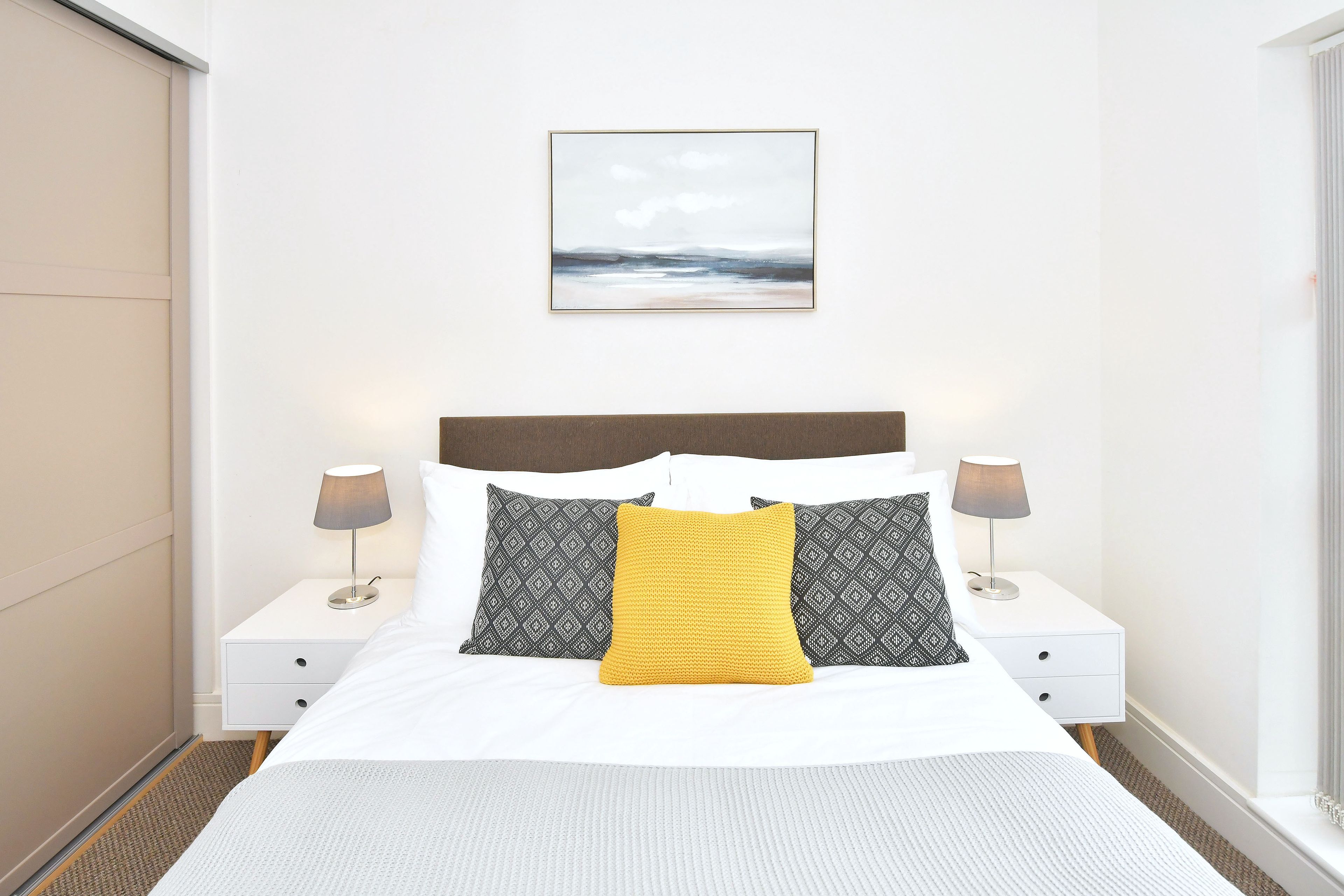 A hotel room with a bed, two bedside tables, a nightstand, a wall-mounted picture, and a window.