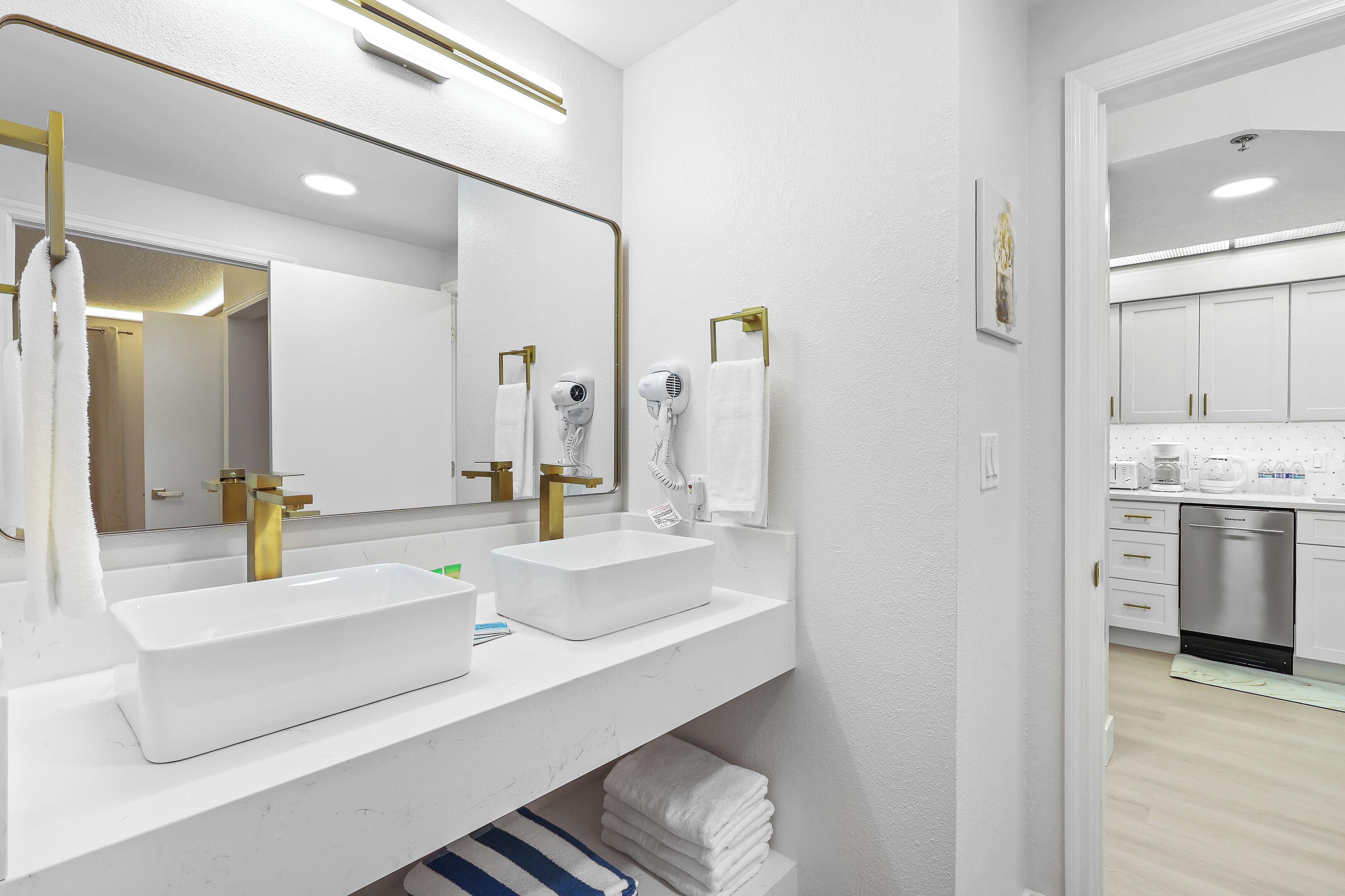 A modern bathroom with two sinks, a large mirror, and a view into the kitchen.