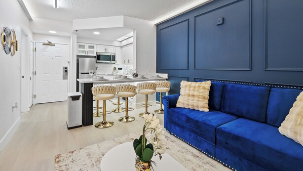 A modern living room with a blue sofa, a white coffee table, and a kitchen area in the background. - 1BR Family Apt, Minutes to Universal & Epic (Orlando)