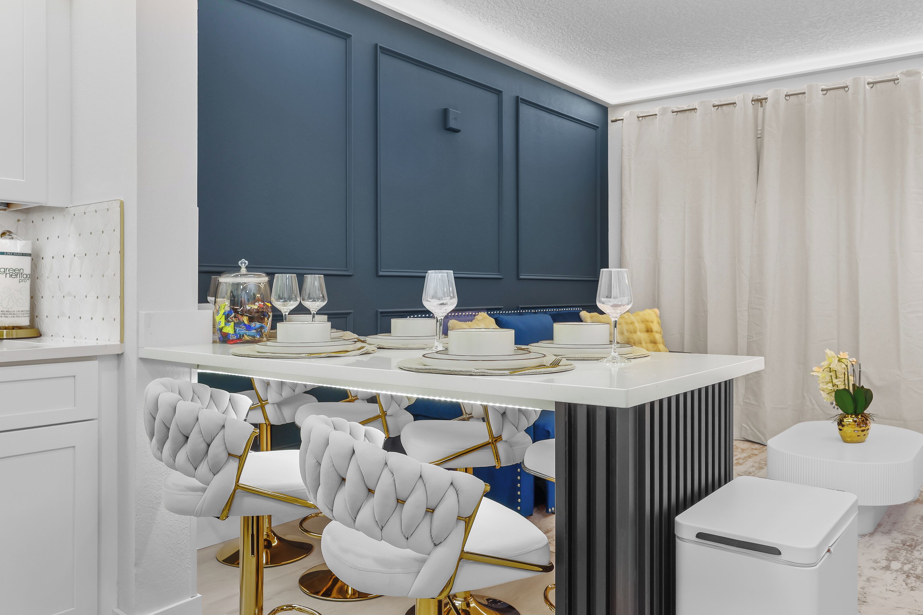 Een moderne eetruimte in een kleurenschema van wit en donkerblauw, met een witte eettafel met gouden accenten, beklede witte stoelen en een witte bijzettafel met een bloemenvaas.