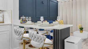 A modern dining area with a white and dark blue color scheme, featuring a white dining table with gold accents, white upholstered chairs, and a white side table with a vase of flowers. - 1BR Family Apt, Minutes to Universal & Epic (Orlando)