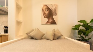 Een keurig opgemaakt bed met een beige sprei en kussens, een aan de muur hangend kunstwerk en een pot met een plant.