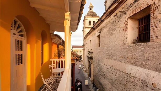 A narrow street with buildings on either side, a balcony with a white chair and table, and a distant tower. - Apt with balcony in the Historic Center - Zaher G (Cartagena)