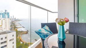 Ein Balkon mit Blick auf Meer und Stadt, eine Vase mit Blumen und ein blaues Kissen.