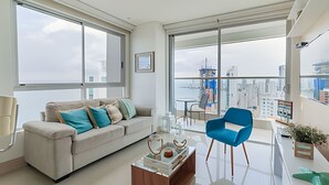 Ein modernes Wohnzimmer mit einer Couch, einem Couchtisch und einem blauen Sessel. Ein Balkon bietet Blick auf die Stadt.