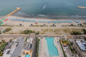 Une vue aérienne d’une plage, avec des rangées de parasols bleus et une piscine située à côté d’une route.
