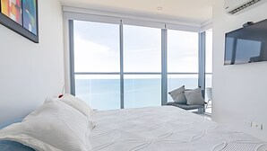 Ein modernes Schlafzimmer mit einem großen Bett, einem Sessel und Blick auf den Himmel durch große Fenster.