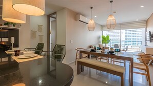 Une cuisine moderne dotée d’un îlot central, d’un coin repas avec des chaises en bois, d’un éclairage suspendu et offrant une vue sur la ville grâce à de grandes fenêtres.