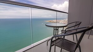 En balkon med gelænder af glas, et rundt bord og to stole, med udsigt over en kystby og havet.