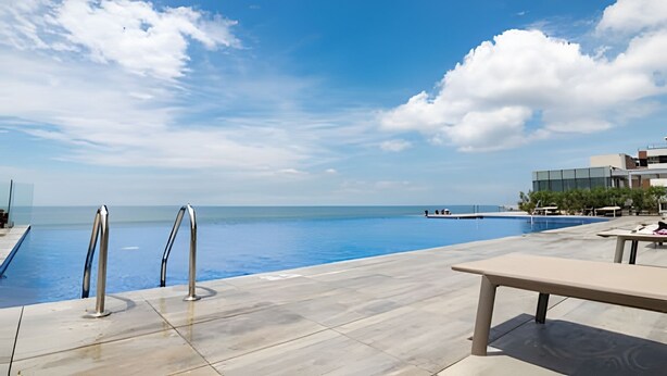 Área da piscina com vista desobstruída para o oceano, uma espreguiçadeira e um edifício moderno ao fundo.