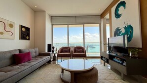 Uma sala de estar moderna com um sofá cinzento, duas poltronas, uma mesa de centro e uma televisão. Janelas amplas oferecem vista para a cidade e para o mar.