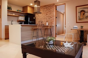 A modern kitchen with a brick wall, wooden bar counter, and bar stools. There is a dining area with a glass table and chairs. - Relaxing New Apt w/ Balcony in Getsemani! (Cartagena)