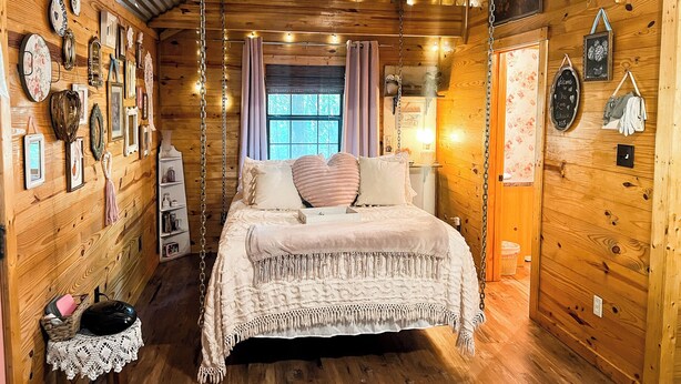 Une chambre confortable, aménagée dans le style d’un chalet, avec un intérieur en bois, un lit recouvert d’une courtepointe blanche et divers éléments de décoration murale, notamment des tableaux encadrés et une pancarte de bienvenue.