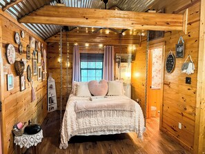 A cozy cabin-style bedroom with a wooden interior, a bed with a white bedspread, and various wall decorations including framed pictures and a welcome sign.