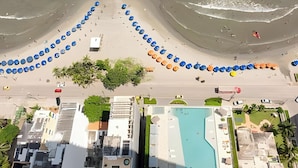 Une vue aérienne d’une plage avec des rangées de parasols et de chaises de plage, une piscine et des bâtiments en arrière-plan.