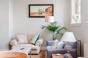 Ein Wohnzimmer mit einer Couch, einem Sessel und einem Tisch mit einer Lampe. An der Wand hängt ein gerahmtes Bild und in der Ecke steht eine Pflanze.