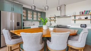 A modern kitchen with teal cabinets, a stainless steel refrigerator, a wooden dining table with four upholstered chairs, pendant lights, and a potted plant. - Stylish Stay w/ All you Need- The Old Grammar School (Kibworth)