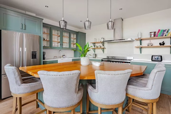 Eine moderne Küche mit türkis-blauen Schränken, einem Edelstahl-Kühlschrank, einem Holzesstisch mit vier gepolsterten Stühlen, Pendelleuchten und einem Blumentopf.