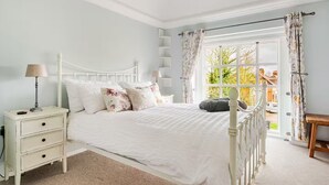 A bedroom with a white bed, a nightstand, a lamp, and a view of the outside through a window with curtains. - Stylish Stay w/ All you Need- The Old Grammar School (Kibworth)