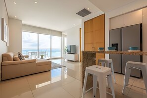 A modern living room with a large window, a sofa, and a kitchen area with stools and a TV. - Spectacular Bay View! 26th Floor H2 Hyatt (Cartagena)