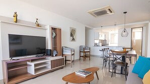 Uma sala de estar moderna com televisão de tela plana, um móvel de entretenimento de madeira, um sofá, uma mesa de centro e um espaço de jantar com cadeiras e uma mesa.