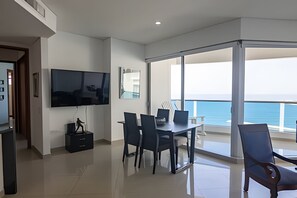 A modern living room with a flat-screen TV, a dining table with chairs, and a balcony with a view of the sea. - Spectacular Ocean View - Infinito Building 1802 (Cartagena)