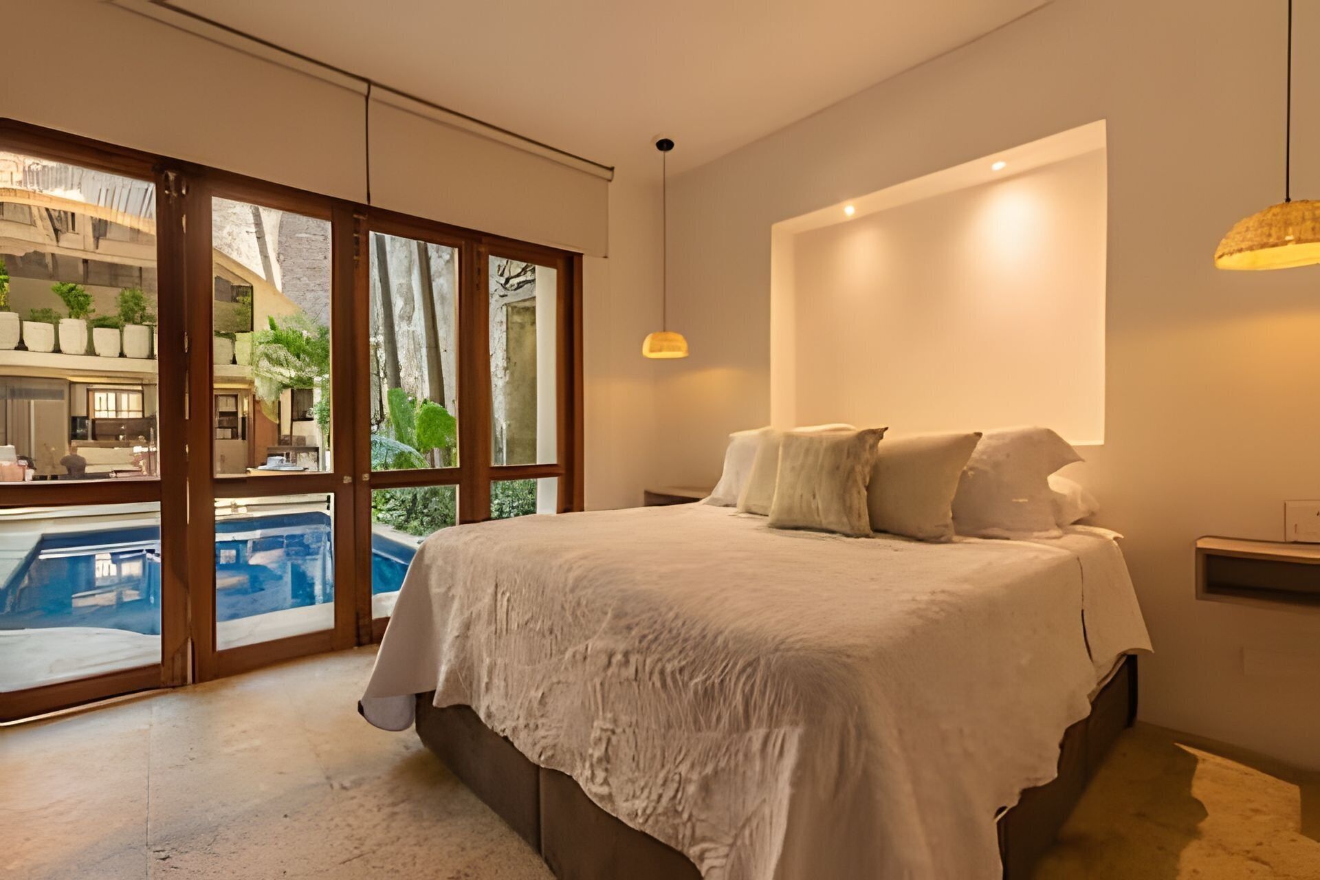A bedroom with a large bed, a view of a swimming pool, and a balcony with plants.