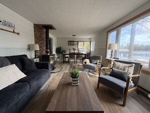 A cozy living room with a fireplace, a dining area, and large windows offering a view of the outdoors. - Sully Shores - Haliburton Lake Getaway (Dysart Et Al)