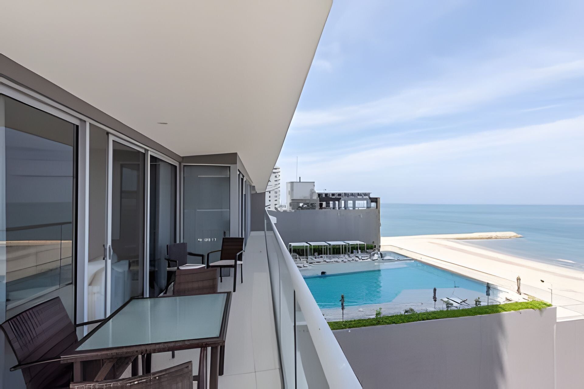 Un balcon avec une table et des chaises en verre, donnant sur une piscine et une plage.