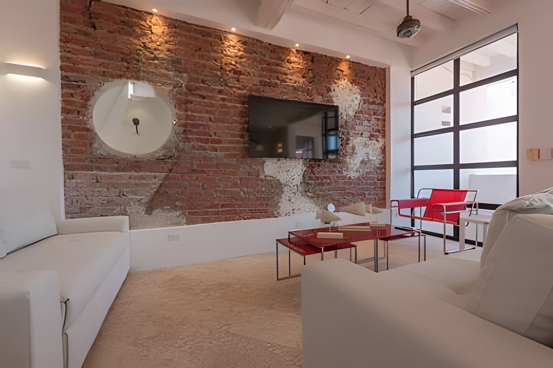 Una sala de estar moderna con pared de ladrillo, televisor montado, mobiliario blanco y un ventanal grande.