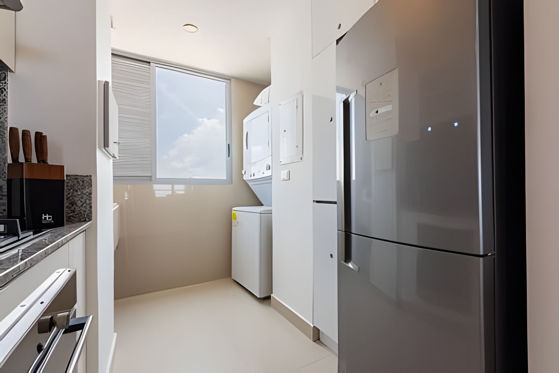 A modern kitchen with a stainless steel refrigerator, white cabinetry, and a window with blinds.