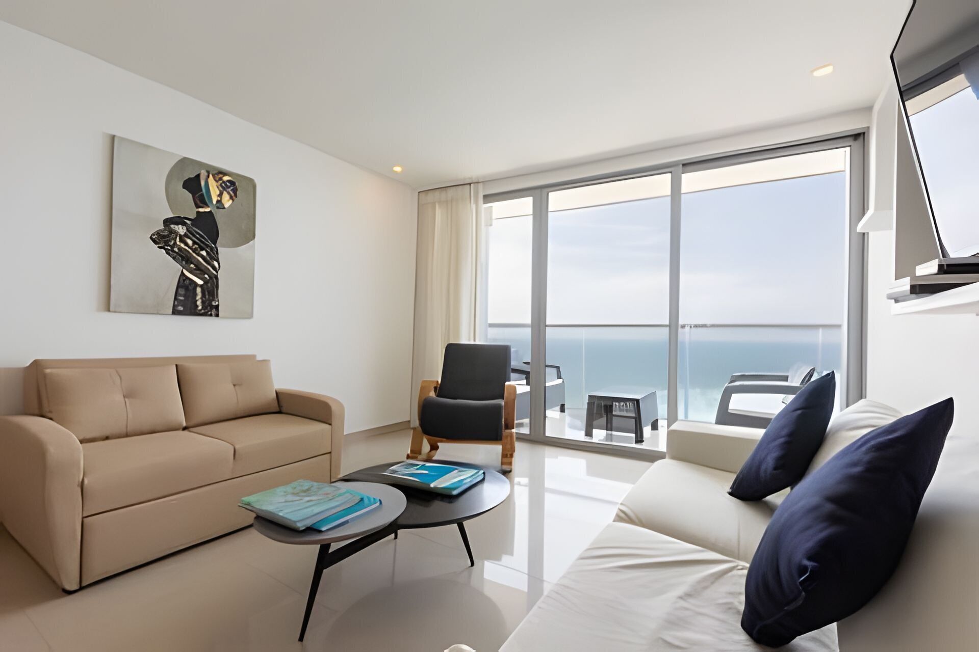 Un salon moderne avec un canapé beige, un fauteuil noir et un canapé blanc agrémenté de coussins bleus. Une table basse ronde est surmontée de livres. Une grande fenêtre offre une vue sur la mer.