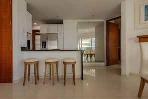 A modern kitchen with white cabinets, a central island with stools, and a dining area visible through an open door. - Spectacular Ocean View - Ed. Infinito 2302 (Cartagena)