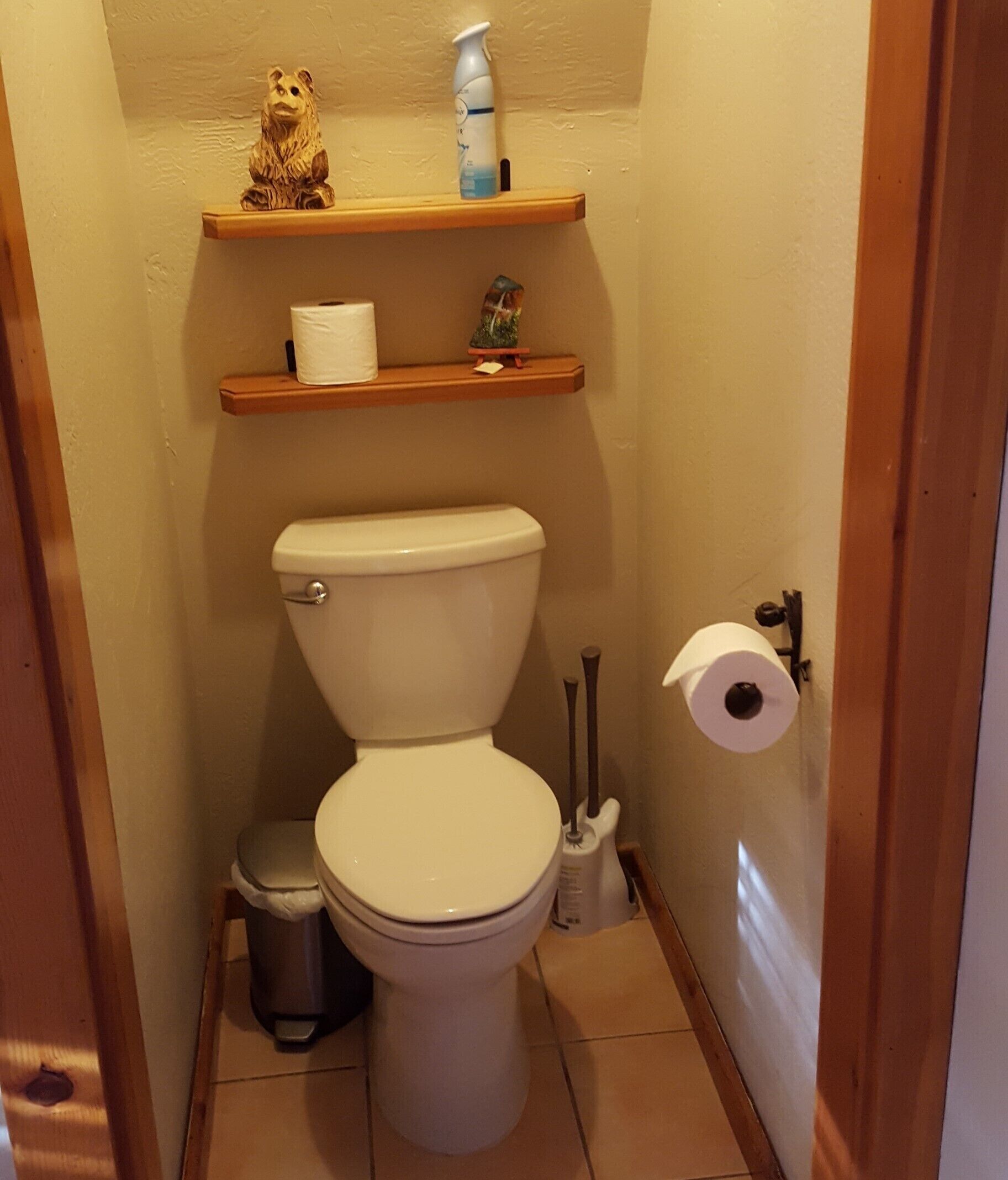 A small bathroom with a toilet, a trash can, a shelf with a bear figurine and a bottle, and a roll of toilet paper.