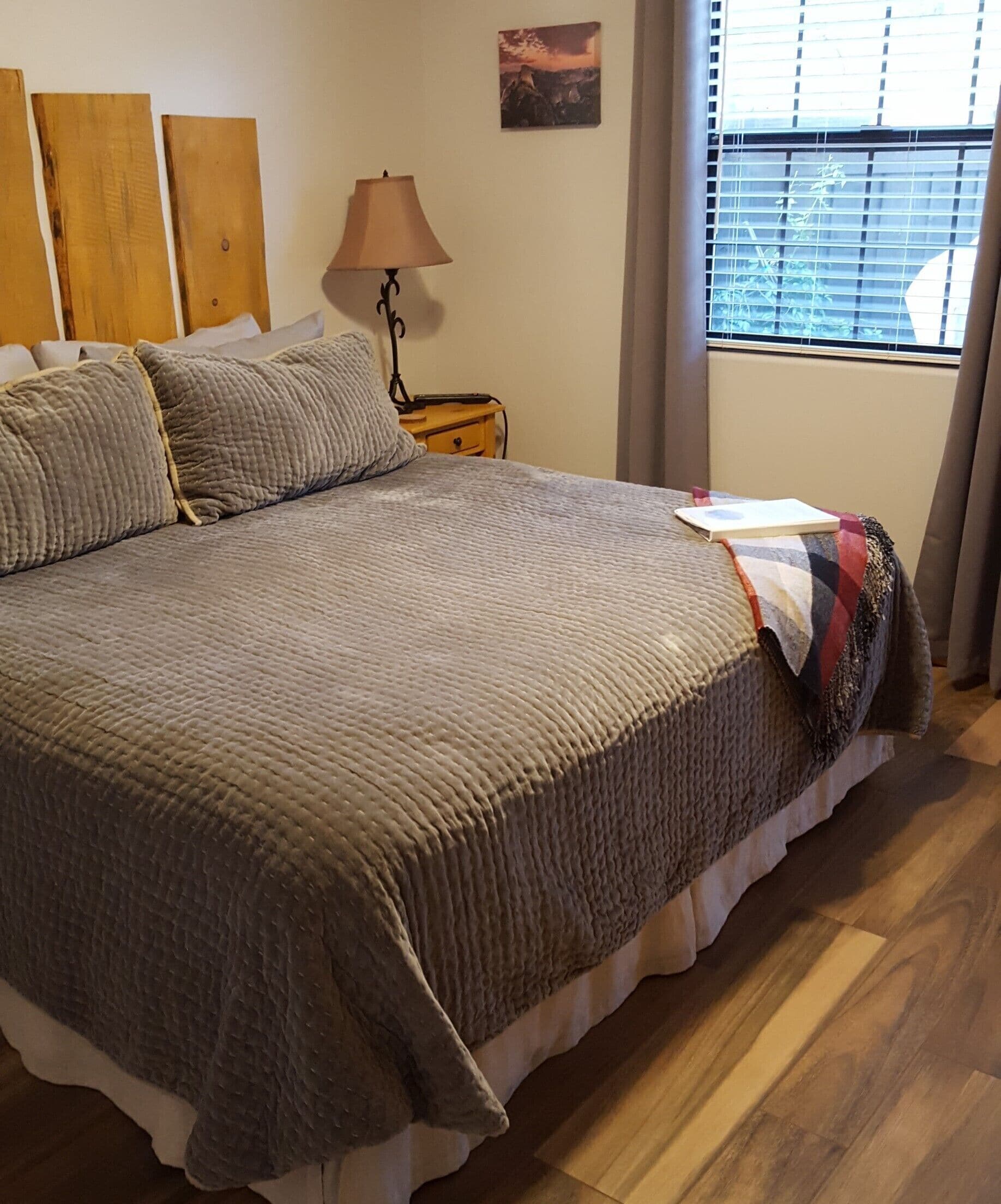 Una cama grande con una colcha texturizada, una lámpara de noche, una mesita de noche de madera y una ventana con persianas.