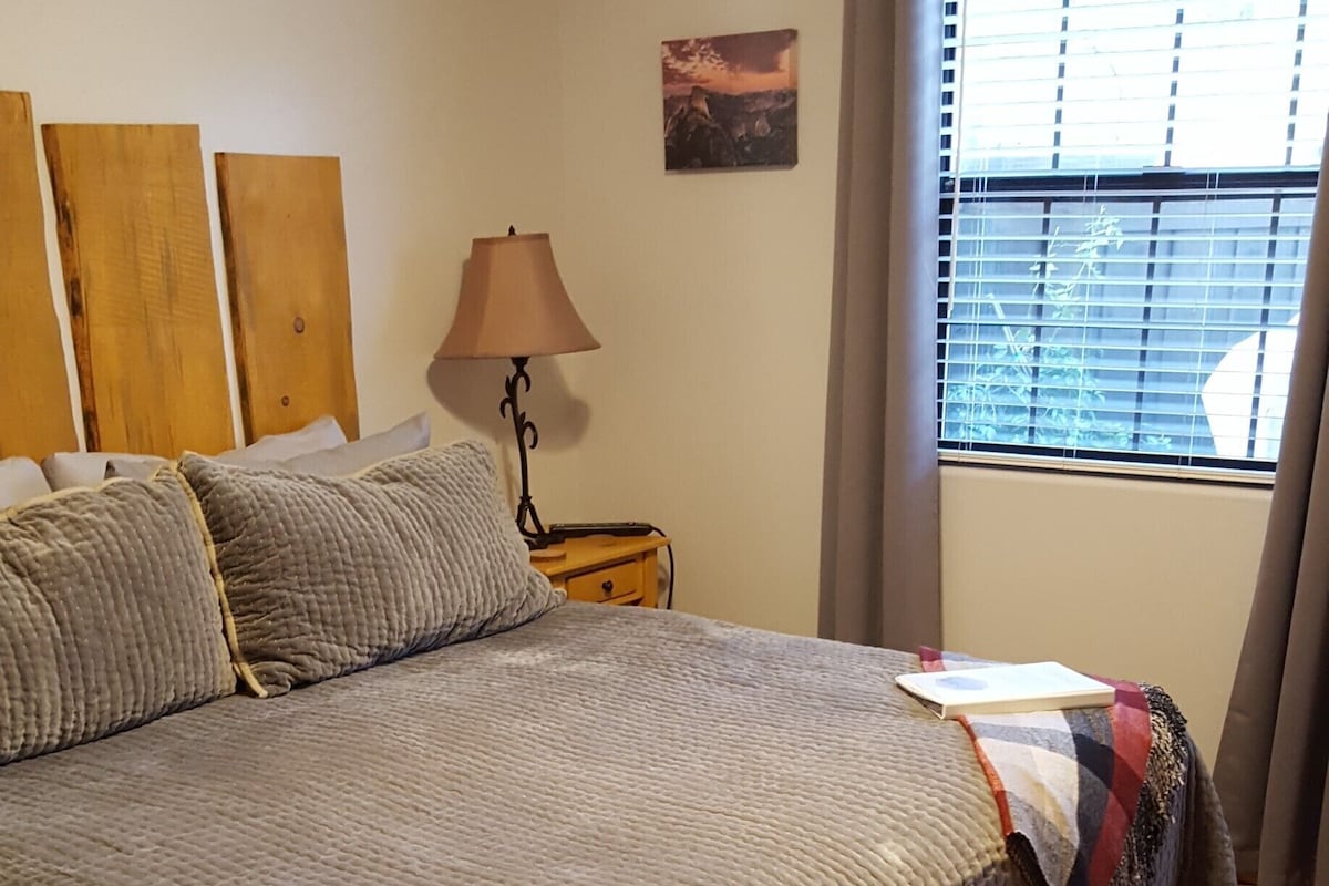 A large bed with a textured blanket, a bedside lamp, a small wooden nightstand, and a window with blinds.