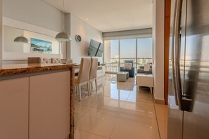 A modern kitchen with a marble countertop, a dining area with white chairs, a flat-screen TV, and a large window offering a city view. - H2 building -Luxurious 2 BR / 16th Fl - Bocagrande (Cartagena)